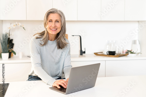 Cheerful mature gray-haired woman spends leisure online, smiling woman sitting in the kitchen with a laptop, watching movies, scrolling news feed in social media, looks at camera and smiles