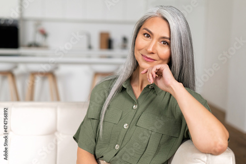 Close-up portrait of charming middle-aged woman with long silver hair sitting in relaxed athmosphere, mature 50s asian female resting at home, looking at camera and smiling with calm smile
