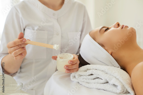 Female cosmetologist stirs and prepares moisturizing cosmetic mask to apply it to client s skin. Close up of jar with white mask or cream in hands of beautician sitting near head of female client.