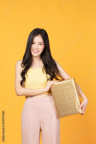 Portrait of happy smiling girl in casual holding gift box and expression face isolated over yellow background