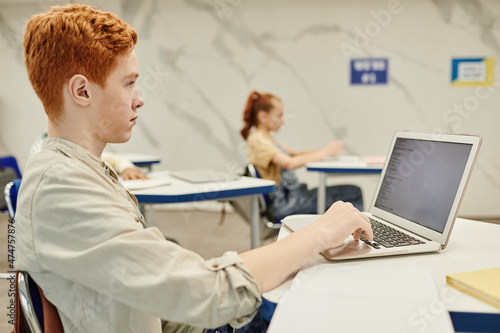 Side view portrait of red haired teenage boy using laptop while coding in school classroom, copy space