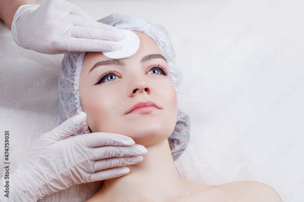 Close up of hands of skillful beautician cleaning and touching female face with cotton pad or sponge. The woman is relaxing.Woman Getting Facial Treatment in Beauty Salon. Close up of hands of skillful beautician cleaning and touching female face with cotton pad or sponge. The woman is relaxing.Woman Getting Facial Treatment in Beauty Salon.