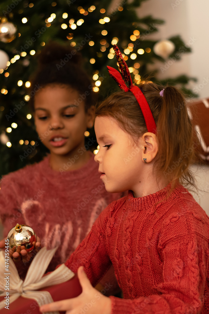 Two sisters in an interracial family unpacks gift boxes at home by the Christmas tree.The girls are dressed in red sweaters and jeans.New Year and Christmas concept.Selective focus. Two sisters in an interracial family unpacks gift boxes at home by the Christmas tree.The girls are dressed in red sweaters and jeans.New Year and Christmas concept.Selective focus.