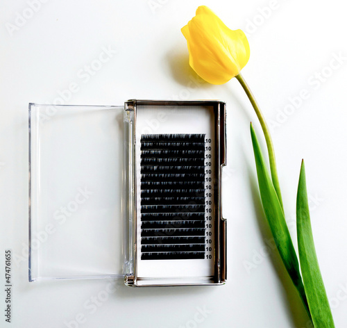 Eyelash extensions in a palette next to a yellow tulip on a white background. Artificial false eyelashes next to yellow flower #474761655