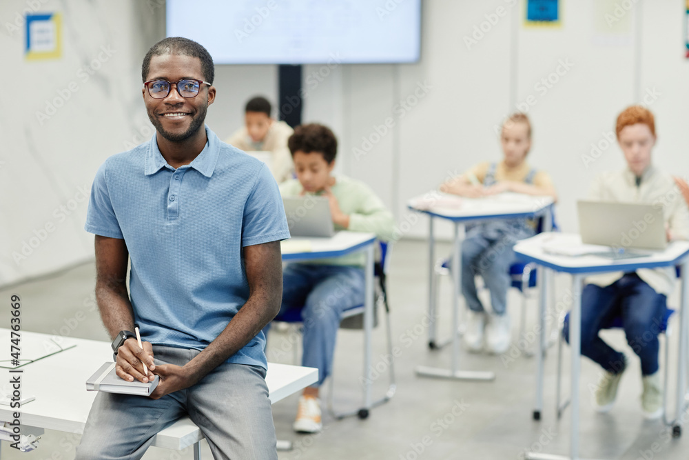 Obraz premium Portrait of young African-American teacher sitting on desk and smiling at camera with children in background, copy space