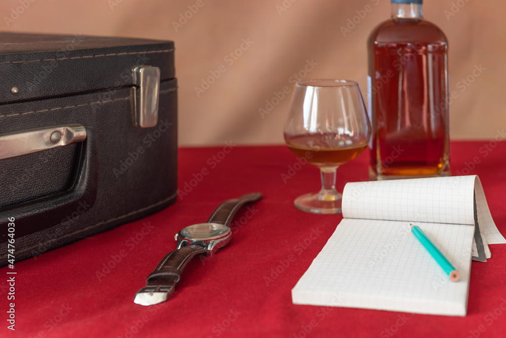 Bottle, glass, wrist watch, notebook, pencil, suitcase. Red tablecloth, brown background. retro Bottle, glass, wrist watch, notebook, pencil, suitcase. Red tablecloth, brown background. retro