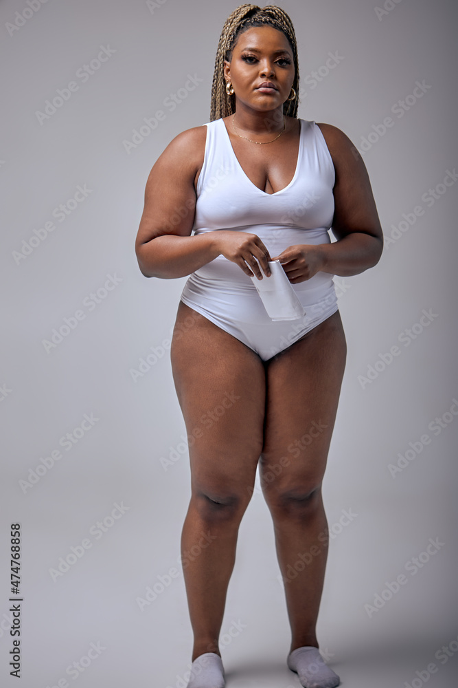 Foto de Young pretty plus size or plump woman with chubby natural body  holding napkin, posing, tired after photoshoot. beautiful female model in  white bodysuit against gray background. Lifestyle portrait do Stock