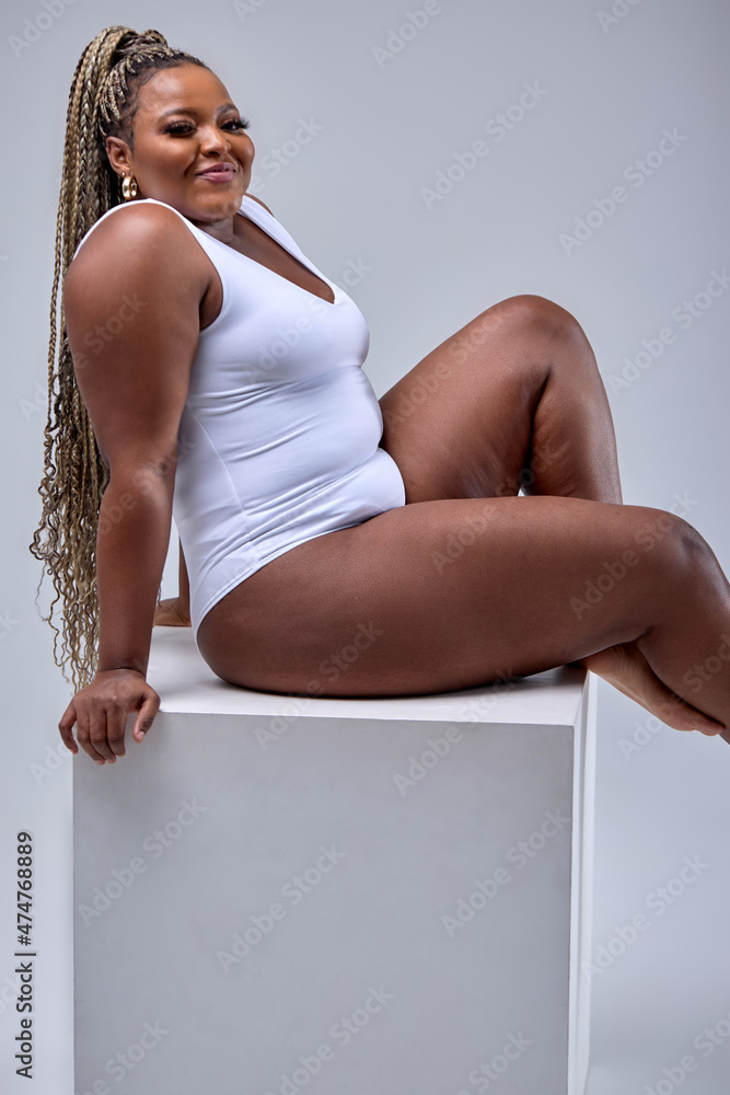 Chubby happy black woman in white bodysuit lingerie posing on white studio  background alone. Plus-size beautiful female model lady. Fat woman. Pin-up  style photo. Copy space to advertise Stock Photo