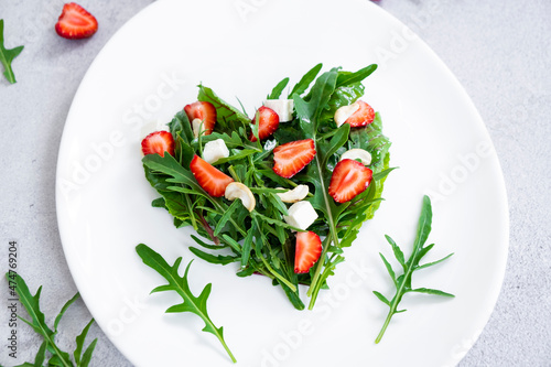Delicious salad of strawberry with mix of herbs, arugula, spinach leaves, feta cheese and cashew nuts in heart shape on white plate. Vegetarian food and raw food diet.