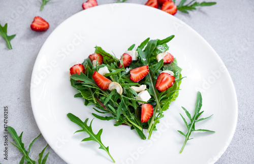 Vegan salad of strawberry with mix of herbs, arugula, spinach leaves, mozarella cheese and cashew nuts in heart shape on white plate. Clean eating, detox and raw food diet.