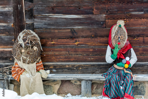 Dolls in Meteņi mumming masks and costumes sitting in a bench near an old wooden house. Meteni is ancient Latvian spring waiting holiday, when mummers scare the winter.