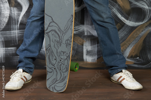 Legs of a fashionable man in jeans and sneakers with a skateboard in his hands close-up. Man poses against a painted wall