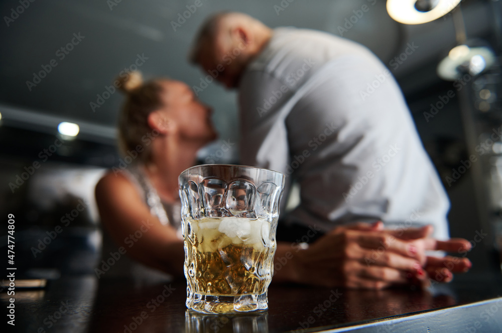 Focus on a glass of alcoholic drink and ice cubes against a blurred background of a middle aged European heterosexual couple flirting together at a bar.