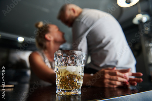 Wallpaper Mural Focus on a glass of alcoholic drink and ice cubes against a blurred background of a middle aged European heterosexual couple flirting together at a bar. Torontodigital.ca
