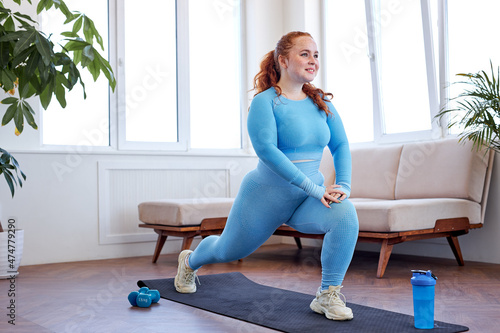 overweight freckled woman doing stretching exercise at home, on fitness mat. attractive caucasian lady in blue sportswear is engaged in sport, preparing for yoga, workout training in living room