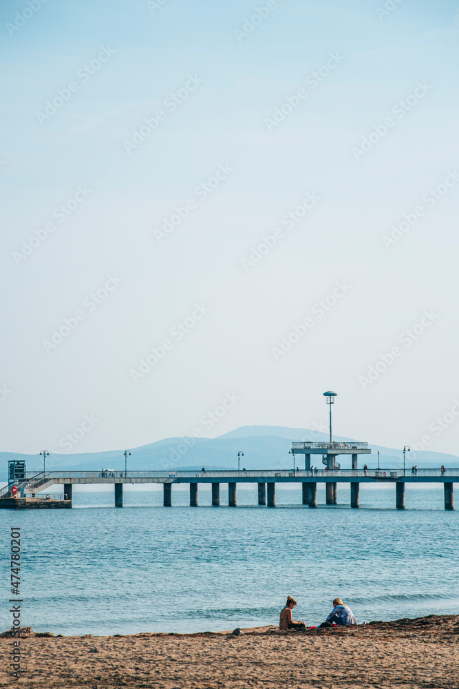 The sea bridge of Burgas, Bulgaria. Blue water and sky in autumn season. Couple sits relaxes on the beach sand.
