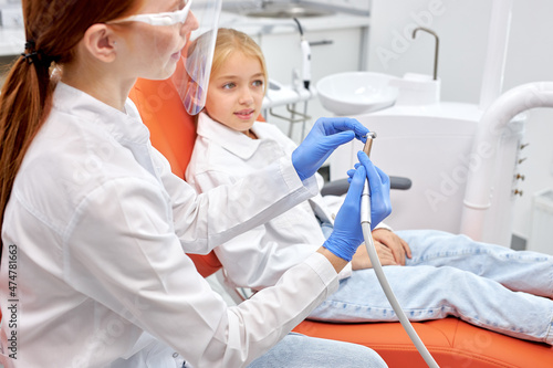 Healthy teeth and smile. Female doctor sitting at chair  holding dental drill in hand. Side view on young woman in white medical uniform going to treat teeth of child girl sitting on dental chair