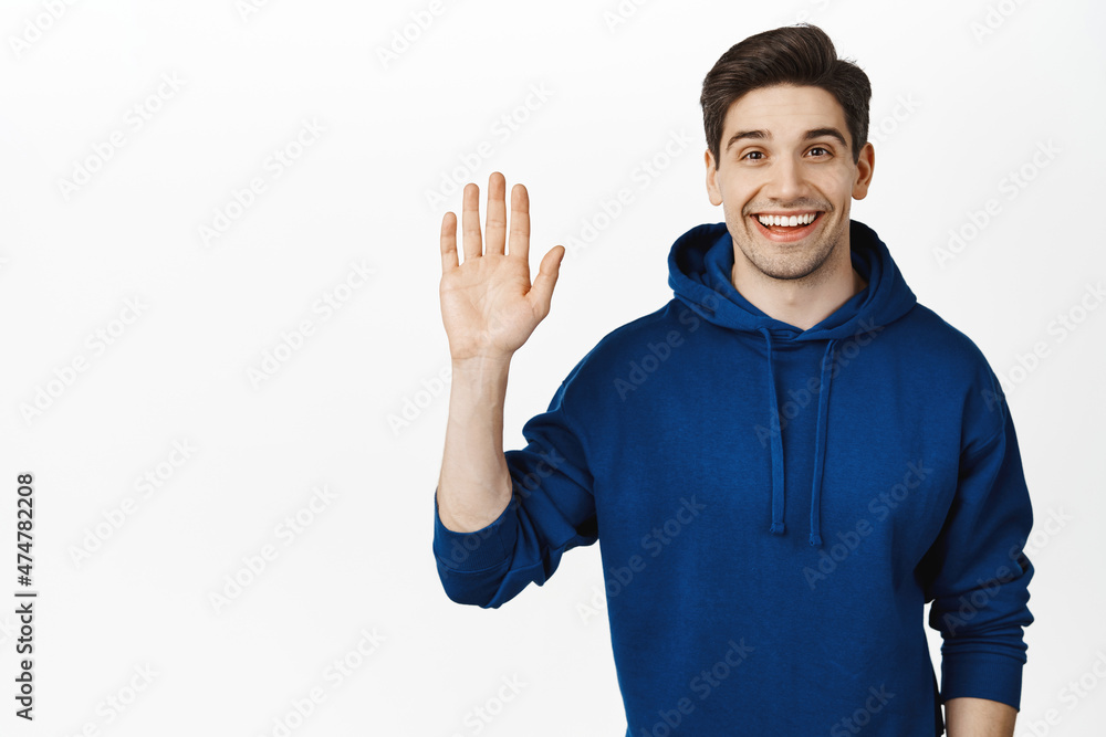 Friendly smiling man waves hand, says hi, hello gesture, greeting guests or customers, standing over white background Friendly smiling man waves hand, says hi, hello gesture, greeting guests or customers, standing over white background