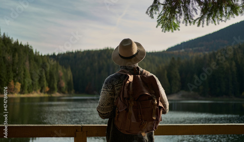 Rear view traveler man wearing hat with backpack standing by mountain lake with calm blue water, enjoying amazing landscape with forest and mountains © vitaliymateha
