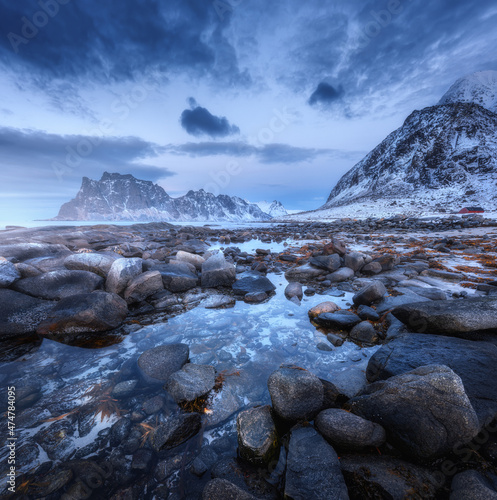 Seashore with stones and blurred water  against snowy mountains and blue sky with clouds at dusk. Uttakleiv beach in Lofoten islands  Norway. Winter landscape with sea  waves  rocks at night. Nature