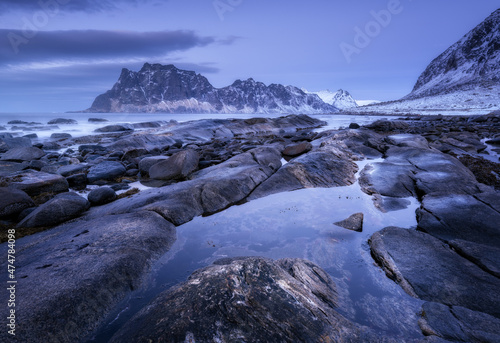 Rocky beach in winter. Seashore with stones, blurred water, snowy mountains and violet sky with clouds at dusk. Uttakleiv beach in Lofoten islands, Norway. Landscape with sea, rocks at night. Nature