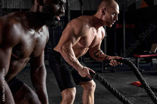 two Athletic young men with battle rope doing exercise in functional training fitness gym. shirtless muscular males holding battle ropes in hands, exercising. side view portrait. cross fit concept