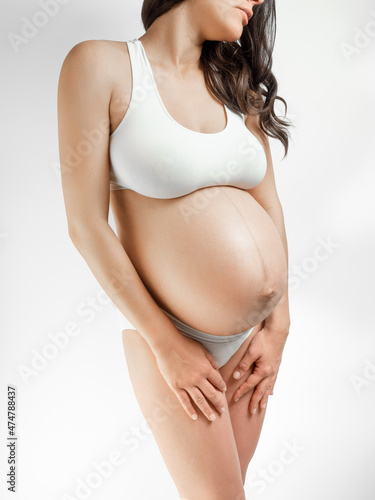 A woman in the third trimester of pregnancy. A pregnant young woman with dark hair stands holds hands in the lower belly against a white background. The girl in the last month of pregnancy in white