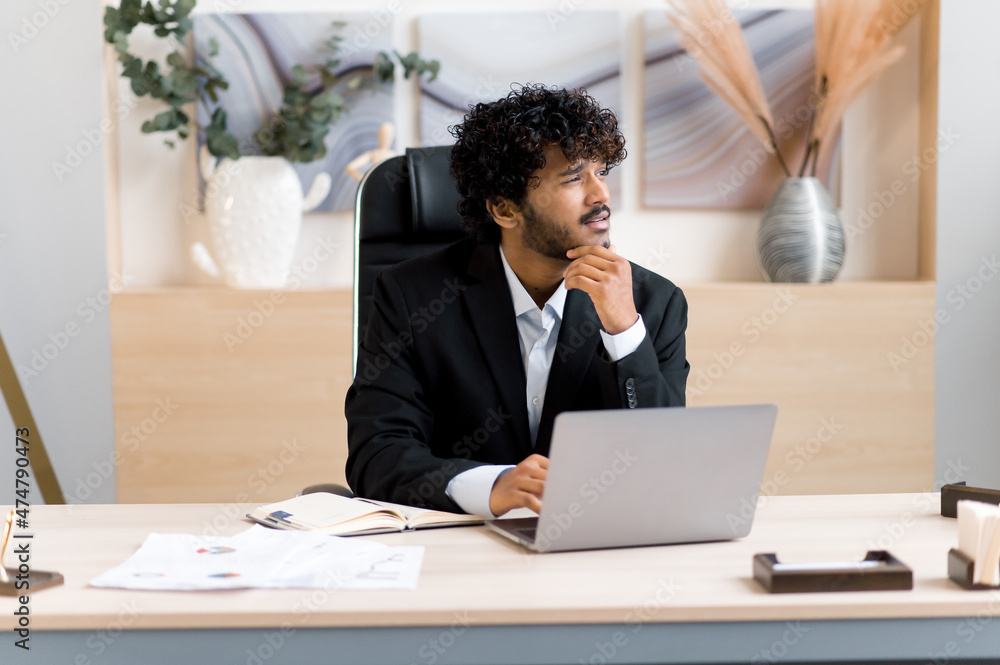 Pensive young successful curly-haired Indian ceo, top manager or businessman in formal suit sitting at table in modern office, look away, thinking over strategy, brainstorming, dreaming about vacation Pensive young successful curly-haired Indian ceo, top manager or businessman in formal suit sitting at table in modern office, look away, thinking over strategy, brainstorming, dreaming about vacation