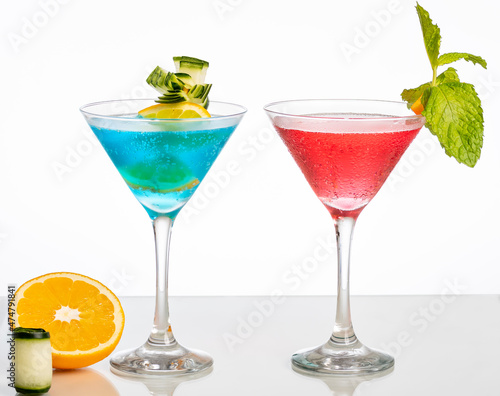 one glass of blue cocktail and one with red cocktail, on reflective surface, isolated on white background.