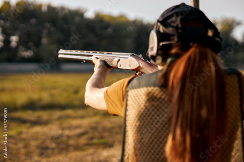 Rear view redhead caucasian woman on tactical gun training classes. Woman with weapon. Outdoor Shooting Range. Side view on beautiful lady with rifle machine gun in the forest, slim female is hunting
