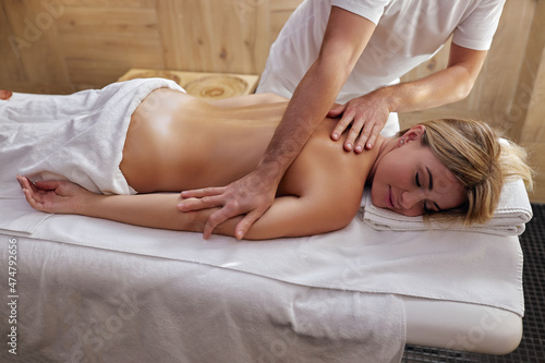 masseur massaging back of young woman lying on massage table at light spa salon. Professional physiotherapist performing wellness treatments for naked slender blonde caucasian female