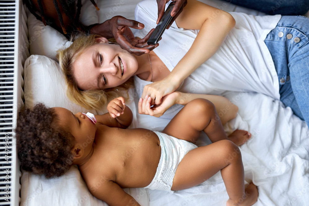 Top View On Interracial Family Having Rest On Bed Using Smartphone, Mobile Phone.View From Above On Black Child And Adult Man With Blonde Lady, Toddler in Domestic Clothes In Cozy Bedroom, Laughing