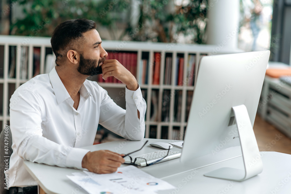 Smart self-confident successful Indian businessman, top manager or ceo with beard sits in an office, wears a white shirt, looks thoughtfully aside, ponders business strategy or project Smart self-confident successful Indian businessman, top manager or ceo with beard sits in an office, wears a white shirt, looks thoughtfully aside, ponders business strategy or project