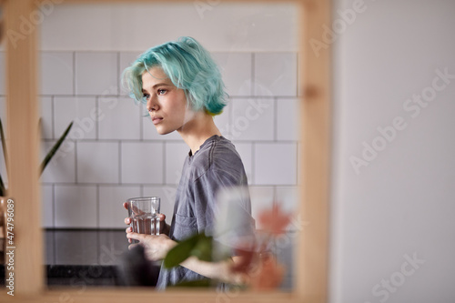 Beautiful caucasian female drinking water in kitchen, alone at home, melancholic mood. attractive lady in casual wear with green hair stand thinking, looking at side in contemplation. loneliness photo