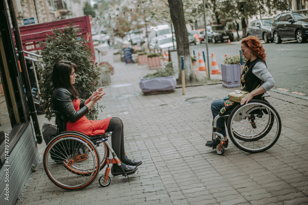Obraz premium Two girls in wheelchairs travel through the central streets. Friends with special needs communicate on the streets of the city