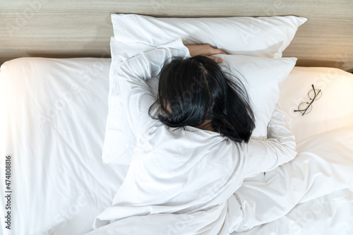 Asian Lady in white long arm t-shirt pajamas lie prone, lie on the stomach in bed with tried and exhausted mood and tone. photo