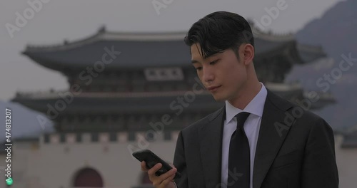 A good looking adult korean office employee dressed in suit texting on his smartphone while waiting patiently for his date in front of historical site of Gyeongbokgung palace, in Seoul, Korea, Asia. photo