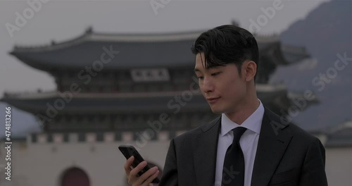 Young asian man pull out his phone and scroll through the notifications on the street with famous heritage building Gyeongbokgung Palace in the background at downtown Seoul city. photo