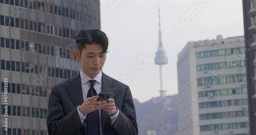 Well groomed korean male office employee in business attire typing messages on his mobile phone with high rise city buildings and YTN Namsam Tower in the background. photo