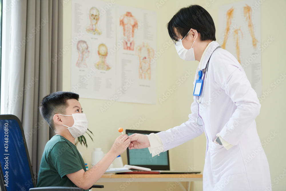 Pediatrician giving lollipop to kid in medical mask afraid of doctors Pediatrician giving lollipop to kid in medical mask afraid of doctors