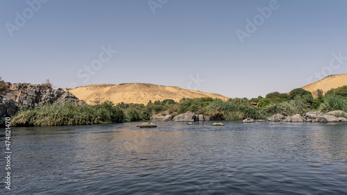 The Nile River flows between the banks with lush green vegetation, picturesque boulders. A sand dune against an azure sky. Ripples and reflections on blue water. Egypt