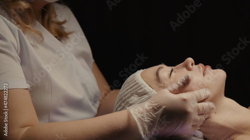 Woman receiving anti-ageing facial massage in spa salon relax. Wellness body skin care face beauty treatment. Black background