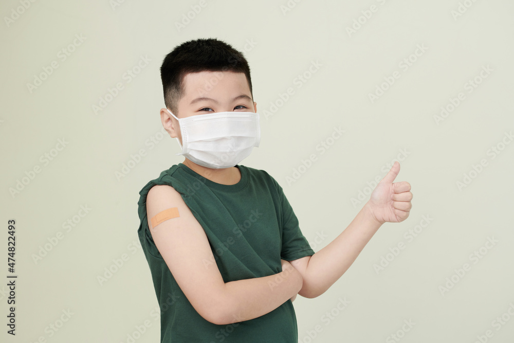 Cheerful little boy wearing medical mask and showing thumbs-up after getting shot of vaccine in arm Cheerful little boy wearing medical mask and showing thumbs-up after getting shot of vaccine in arm