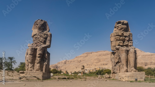 Giant ancient statues of the colossi of Memnon rise in the valley. Huge figures of sitting people are dilapidated. A picturesque sandy mountain against a blue sky. Egypt