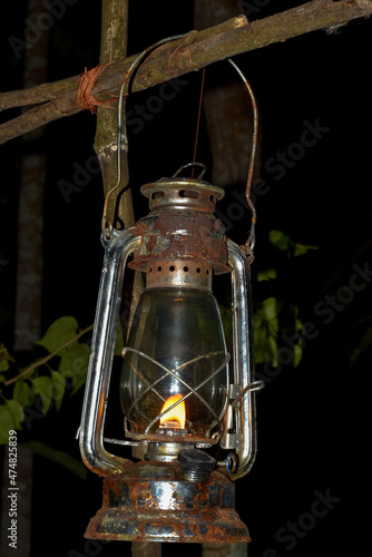 Old kerosene lantern hanging on the dead dry wood for light in forest at night. Travel camping concept.