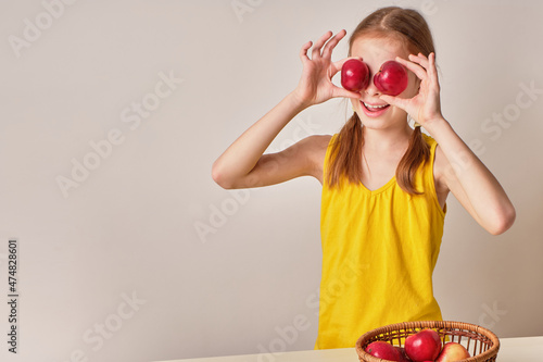 A positive playful girl with apples on a gray-white background. A happy child, a smiling girl holding fresh red ripe apples in her hand. There is a basket of apples nearby. The concept of healthy and
