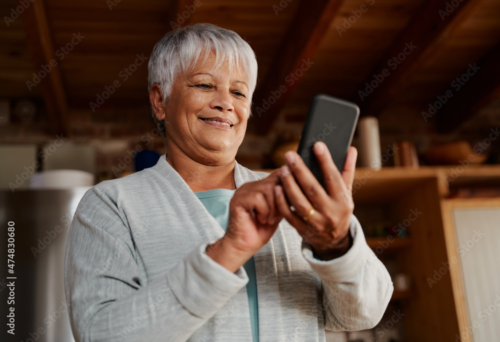 Happy multi-cultural elderly female typing a message on smartphone while standing in modern kitchen holding morning coffee. Happy multi-cultural elderly female typing a message on smartphone while standing in modern kitchen holding morning coffee.