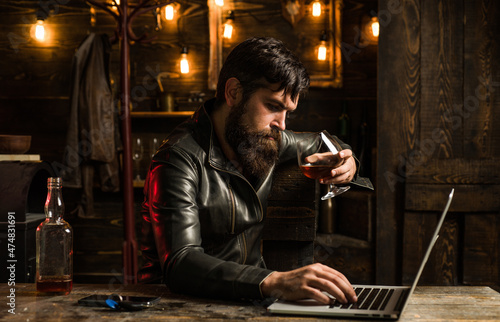 Businessman stressed with wiskey. Handsome man with a beard is sitting in a cafe with glass of whiskey and a laptop.