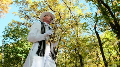 Energetic elderly man plays song with saxophone performing in sunny autumn city park against lush yellow trees low angle shot