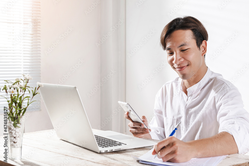 Smart Asian young businessman takes a note from smartphone working at home office pleased happy cheerful on online meeting video or learning by a laptop computer for job work or makes good dealing Smart Asian young businessman takes a note from smartphone working at home office pleased happy cheerful on online meeting video or learning by a laptop computer for job work or makes good dealing